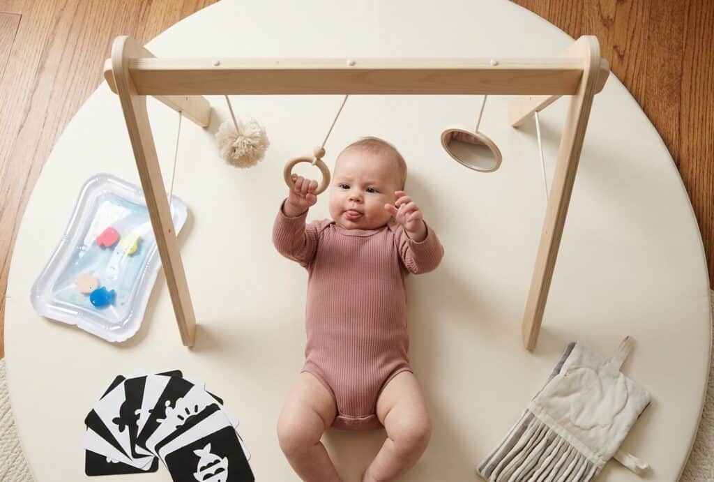 Overhead view of a baby lying on a circular cream play mat reaching up to bat at hanging toys on a minimalist wooden play gym with a water mat and high-contrast art cards arranged nearby on hardwood floor