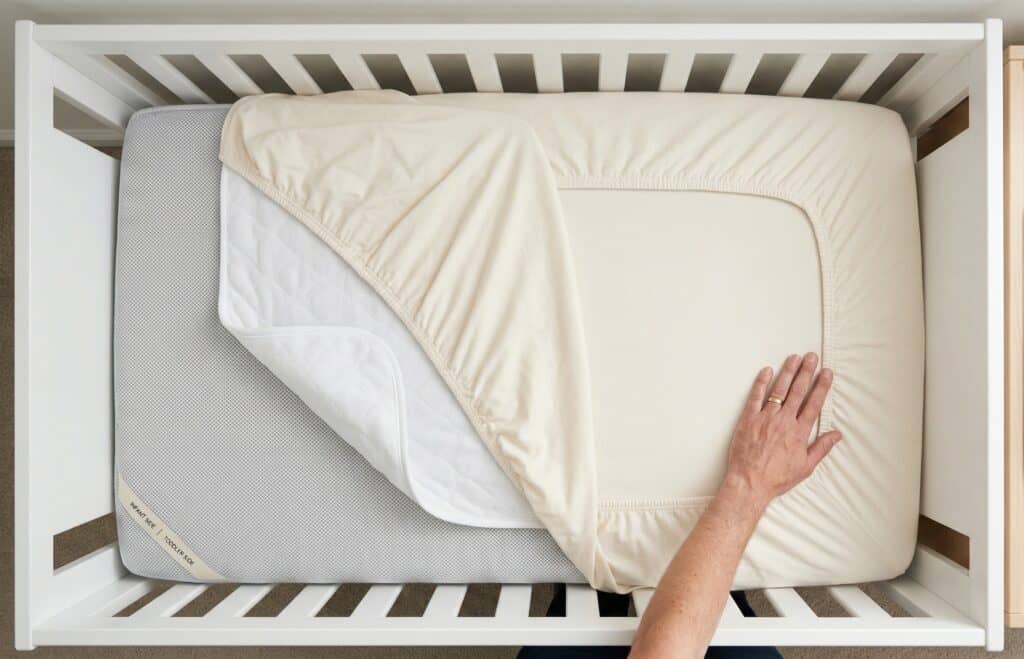 Overhead view looking down into a white crib showing separated bedding layers including a breathable mesh mattress cover, waterproof protector, and tight-fitting organic cotton sheet with a parent's hand smoothing the elastic edge