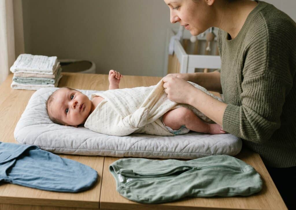 Mother mid-swaddle wrapping her newborn in a cream muslin blanket on a changing table with a velcro swaddle and a zip-up swaddle pod laid out beside the baby for comparison in soft morning light